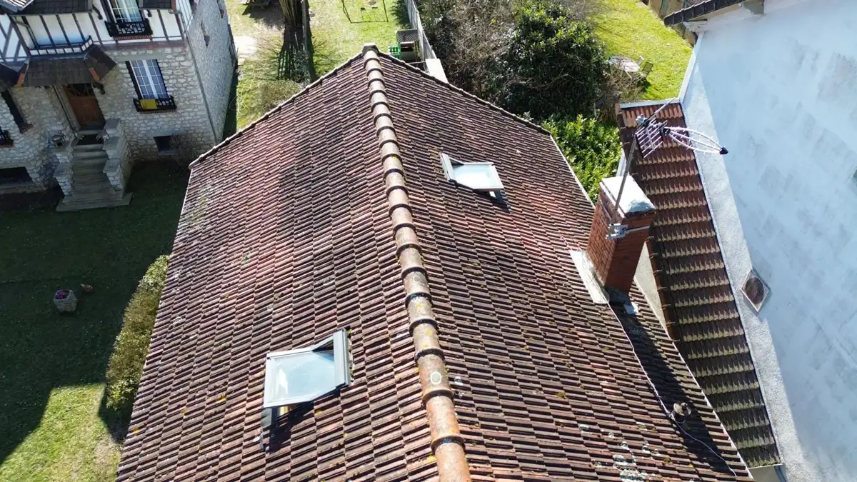 Vue de drone d'une toiture en tuiles mécaniques brunes et usées, avec deux fenêtres de toit, avant réfection
